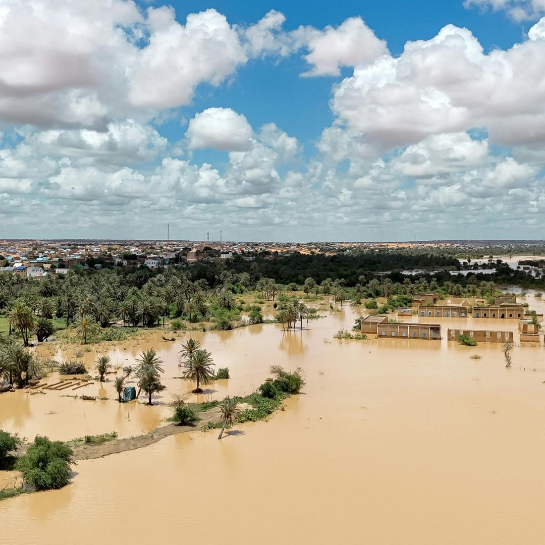 An aerial view of a severe disaster zone, showing vast expanses of muddy brown floodwater completely submerging land, trees, and roads near a populated area.